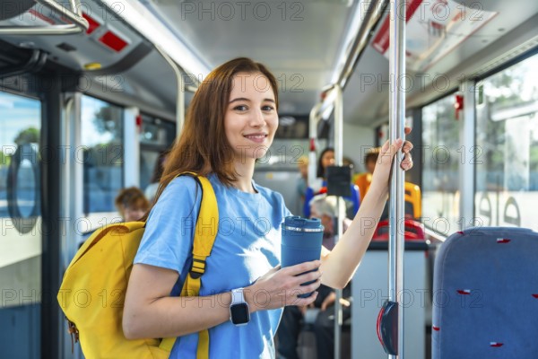 Young woman smiling on a city bus, standing and holding a pole with a yellow backpack and reusable cup, eco conscious commuter enjoying a daytime urban journey