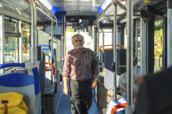Senior man with a beard and plaid shirt standing in the aisle of a public bus, commuting during the day, with other passengers seated and windows bringing in natural light