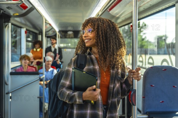 Young black woman with curly hair and glasses smiling, holding a digital tablet and a pole while standing in a public bus filled with diverse passengers, representing urban travel and daily commute