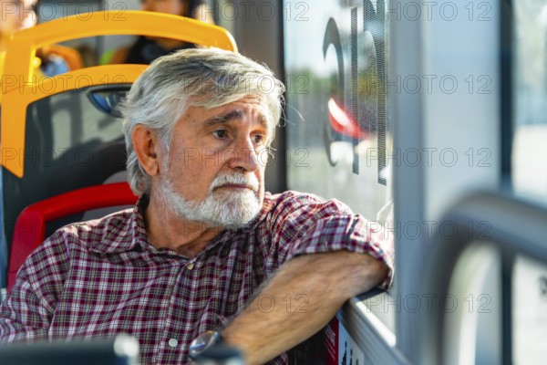 Senior man with gray hair and beard traveling on a public bus, wearing a plaid shirt and looking pensively out of the window, reflecting on his journey