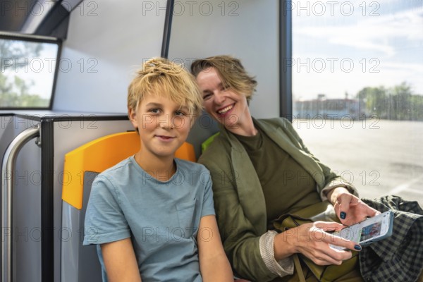 Mother and son sitting inside a bus, sharing a moment while traveling using public transport, the mother scrolling on a smartphone and the son looking at the viewer