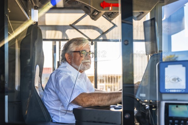 Senior bus driver with eyeglasses and white shirt focused on steering from the cabin, demonstrating dedication and experience in urban public transportation during daytime commute