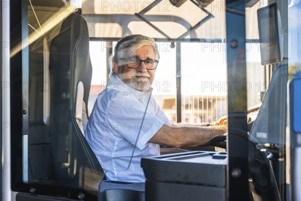 Senior man with a white beard and glasses wearing a shirt, sitting at the driver's seat of a modern bus during daylight, smiling and working in public transport