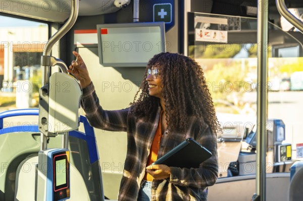 Young black woman holding a digital tablet and validating her public transport ticket on a modern bus, smiling as she is commuting and traveling by urban transit