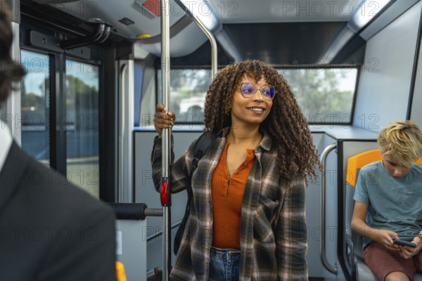 Woman with afro hair and eyeglasses standing and holding a handrail on a public transport bus, smiling while traveling, engaging in daily commute or urban journey