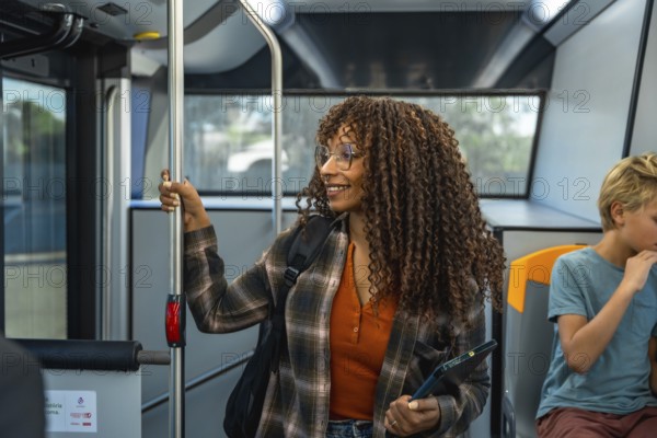 Woman with glasses and curly hair holding a digital tablet while commuting on a public bus, smiling and holding a handrail, with another passenger sitting nearby