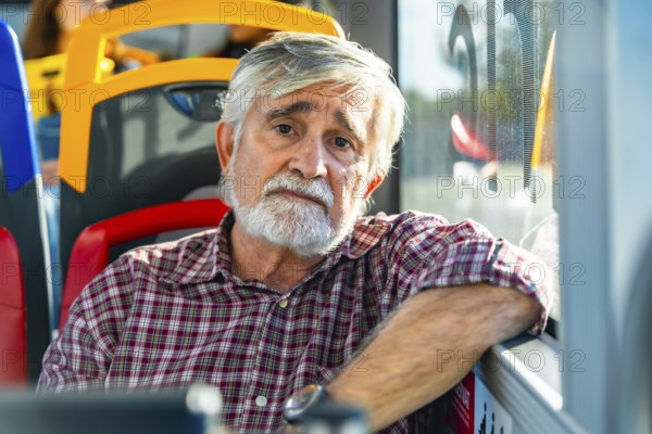 Senior man with white beard and plaid shirt sits by a bus window, gazing pensively at the camera during an urban commute, conveying quiet reflection and daily routine