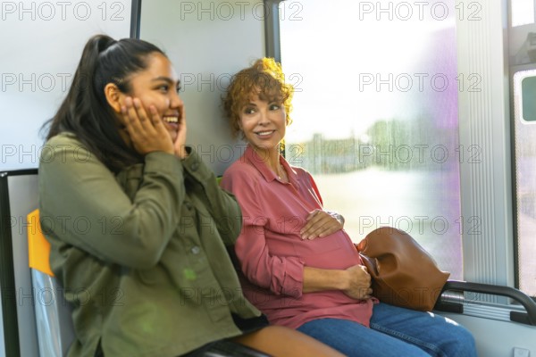 Young woman is smiling and showing surprise while looking at her pregnant friend, who is gently cradling her baby bump, both traveling together on a public bus