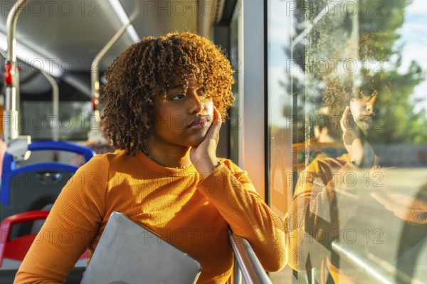 Young woman with curly hair riding public transport bus, holding a laptop and contemplating while looking out the window, reflecting a pensive and introspective moment