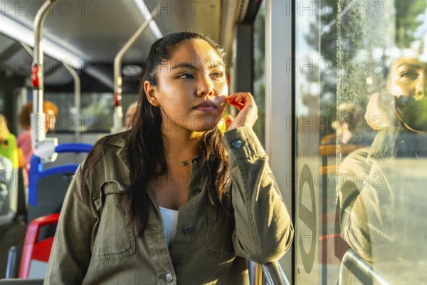 Young woman passenger traveling on a public transport bus, looking out of the window with a pensive expression, enjoying sunlight with her face reflecting on the glass