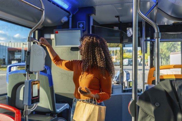 Young woman with curly hair validating her public transport ticket on a bus, holding a smartphone and a paper shopping bag, commuting morning or evening