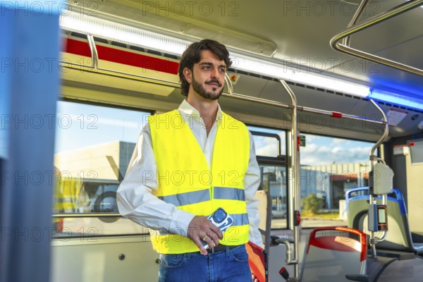Man wearing an unbuttoned white shirt, blue jeans, and a yellow reflective safety vest, holding a phone, and standing confidently in a public transport bus