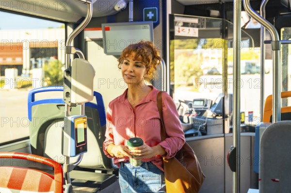 Woman standing on a city bus during the morning commute, holding a reusable coffee cup and brown shoulder bag, smiling and looking forward while traveling through an urban route
