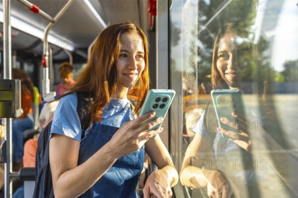 Young woman smiling on a sunny bus commute, holding her smartphone and looking out the window with her reflection in the glass, relaxed and connected during travel