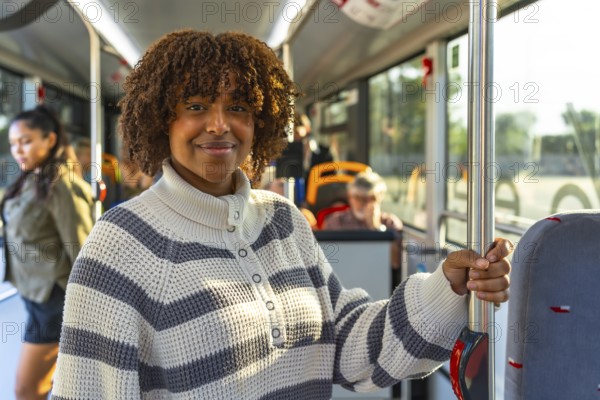 Smiling young woman with curly hair standing on a busy city bus, holding a pole and looking at the camera as sunlight fills the modern interior during her commute