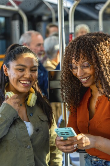 Two young adult women smiling and looking at a smartphone, enjoying public transportation while commuting on a bus with other passengers in the background