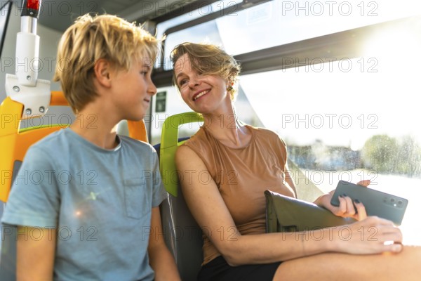 Happy mother and son enjoying a conversation while sharing a seat on a public transport bus, with bright sunlight streaming through the window creating lens flare