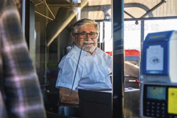 Mature man working as a bus driver, wearing glasses and a white uniform shirt, sitting inside the cabin and smiling at the passenger, representing public service and commitment