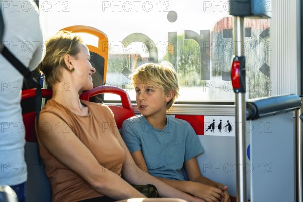 Mother and son smiling and chatting on a sunlit city bus during their commute, sharing a relaxed bonding moment as they travel together through the urban landscape