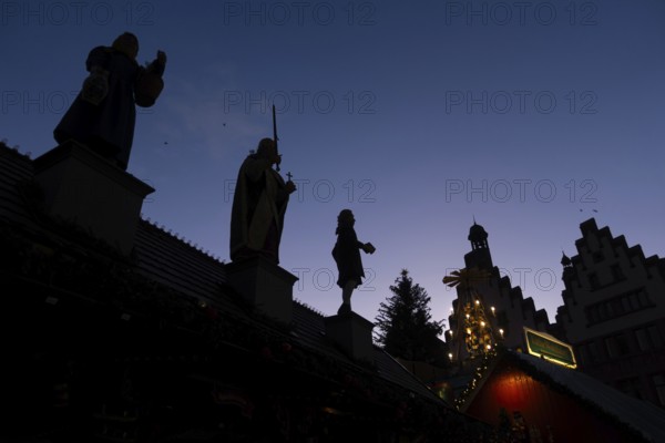The silhouettes of the city greats Mrs. Rauscher (left), Charlemagne (center) and Johann Wolfgang Goethe (right), who are on the roof of a Christmas market stand on the Römerberg in Frankfurt am Main, stand out against the blue evening sky. The Frankfurt Christmas Market starts on November 24 and runs until December 22, Römerberg, Frankfurt am Main, Hesse, Germany