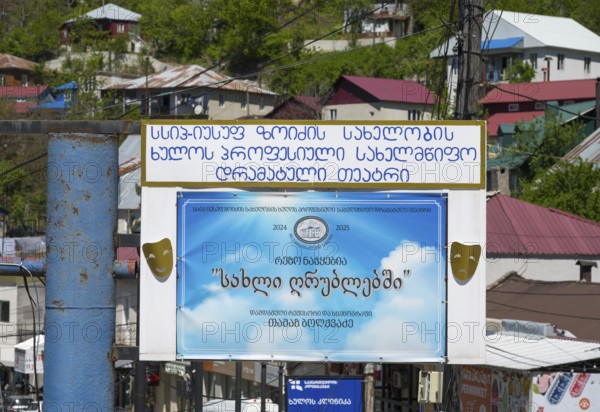 A large advertising sign with a blue poster and text surrounded by colorful buildings and green trees, reference to a theatre, Khulo, Chulo, Autonomous Republic of Adjara, Adjaria, Lesser Caucasus, Georgia