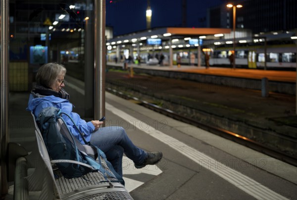 Night shot, elderly woman, traveler, sitting alone on a bench on a platform, backpack, using smartphone, cell phone, main train station, Stuttgart, Baden-Württemberg, Germany