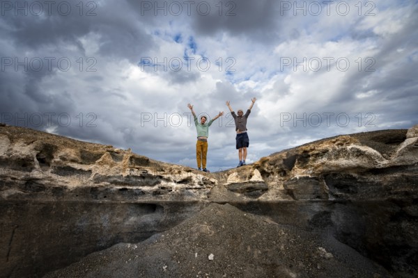 Two young men stretch their arms in the air, Eroded rock formations in volcanic landscape with dramatic cloudy skies, Ciudad Estratificada or Los Roferos, Antigua Rofera de Teseguite, Lanzarote, Canary Islands, Spain