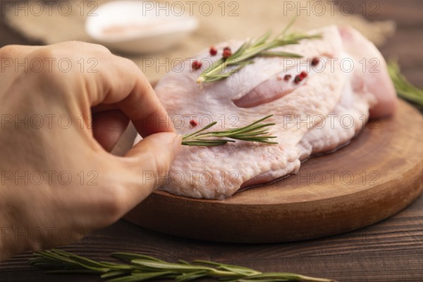 Raw Turkey Thigh with spices and rosemary on cutting board with hand on brown wooden background and linen textile. side view, close up, selective focus