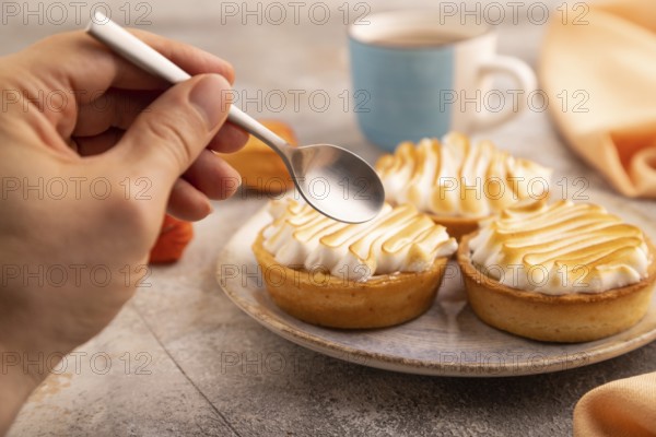 French lemon tart with meringue with hand on brown concrete background, cup of coffee, orange linen textile, side view, close up, selective focus