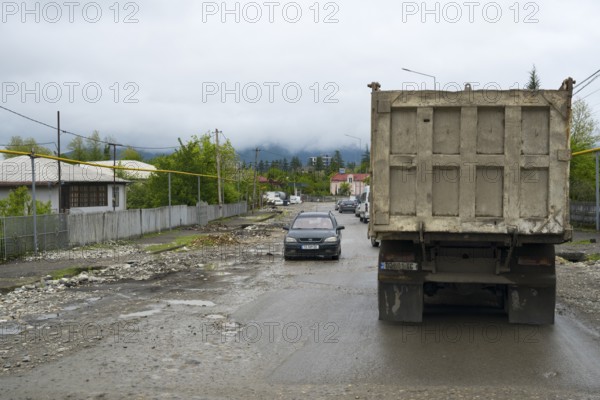 A truck and a car on a wet rural road with cloudy sky and mountains in the background, bad road in a village in Georgia