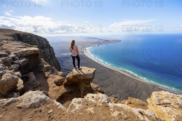 Young woman enjoying the view from the Risco de Famara cliffs to Famara beach, Playa de Famara with La Calaeta, Lanzarote, Canary Islands, Spain