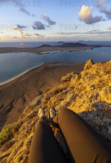 First person view, legs of a woman sitting on a steep cliff, view of the sea and the island of La Graciosa, Mirador del Porrito viewpoint at sunset, Lanzarote, Canary Islands, Spain