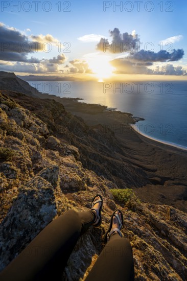 First person view, legs of a woman sitting on a steep cliff, view of sea and coast, Mirador del Porrito viewpoint at sunset, Lanzarote, Canary Islands, Spain