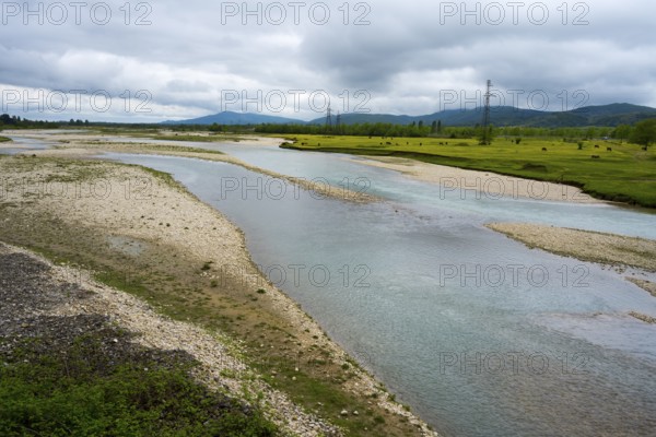 A calm river flows through a pebble landscape under a cloudy sky with power lines in the background, river Abasha or Abashistskali or Abashatskari, near Bandza, Samegrelo-Zemo Svaneti region, Georgia