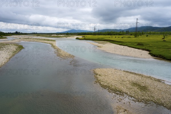 A river snakes through strips of pebbles flanked by green fields and cloudy skies, interrupted by power poles, river Abasha or Abashistskali or Abashatskari, near Bandza, Samegrelo-Zemo Svaneti region, Georgia