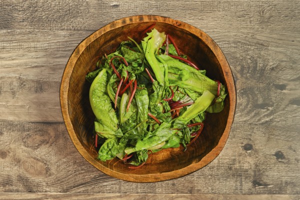 A wooden bowl filled with steamed greens on a rustic wooden tabletop. The leafy vegetables, including romaine and other greens, look crisp and healthy, ready for preparation