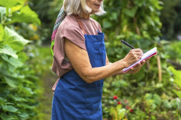 Senior woman in apron and colorful scarf jotting notes in a notepad with a pen while standing in a lush green garden, planning and tracking her gardening tasks and harvest