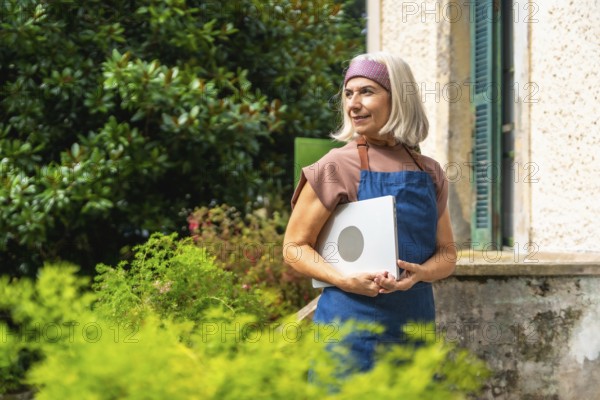 Senior woman in apron and headband holding a laptop outdoors in her garden, smiling and looking away while managing a small business or enjoying remote work in nature