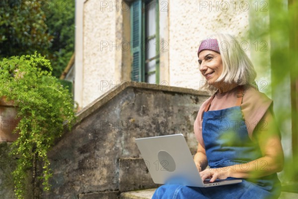 Senior woman enjoying remote work from a peaceful garden setting, smiling while typing on a laptop, embracing a flexible digital nomad lifestyle and retirement