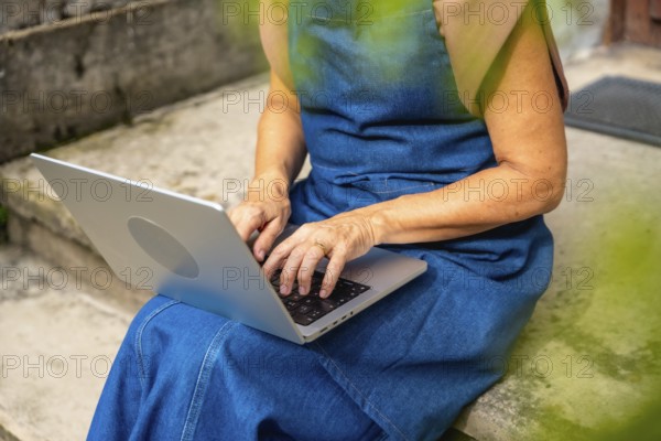 Senior woman on concrete steps typing on laptop outdoors, enjoying flexible remote work and modern connectivity while concentrating on screen in a casual, independent lifestyle setting