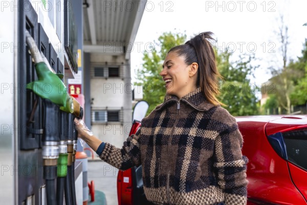 Woman smiling at a gas station while holding a fuel pump nozzle beside her red car in a plaid jacket, preparing to refuel on a sunny day during a road trip stop