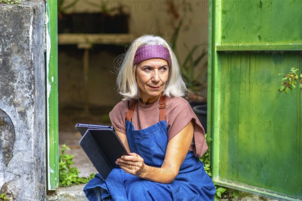 Senior woman artist in an apron and headband working on a digital tablet outside a rustic green building, thoughtful and focused as she sketches and plans her creative project