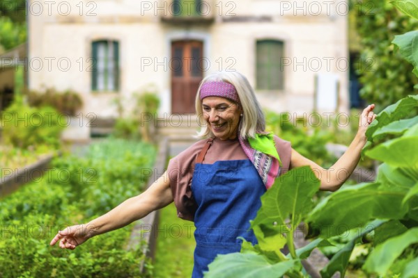 Senior woman with gray hair and colorful headband smiling while standing in her flourishing home vegetable garden, appreciating sustainable and healthy living