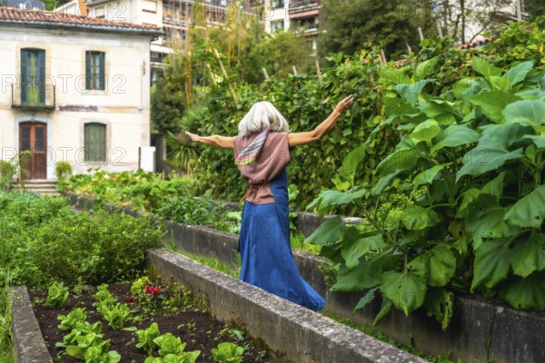 Senior woman with gray hair and scarf feeling freedom and happiness, walking with arms outstretched along a garden path with raised beds filled with plants