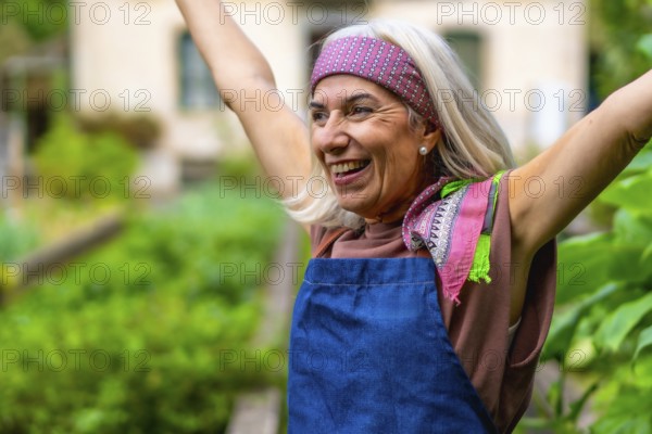 Senior woman in a bandana and apron raises her arms in a sunlit garden, smiling with joy and vitality as she celebrates healthy, active retirement and a flourishing outdoor hobby