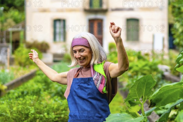 Happy senior woman dancing with arms raised in her home garden, celebrating an active and joyful retirement while cultivating fresh vegetables outdoors