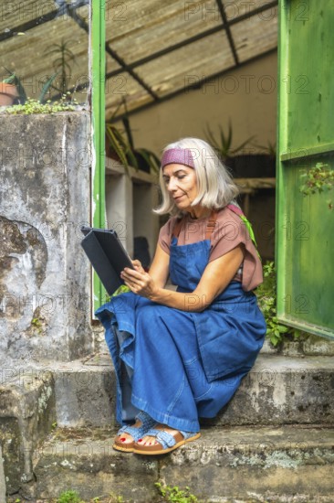 Senior woman in denim overalls and headband sits on rustic outdoor steps in a garden, focused on a digital tablet, enjoying relaxed browsing and modern connectivity