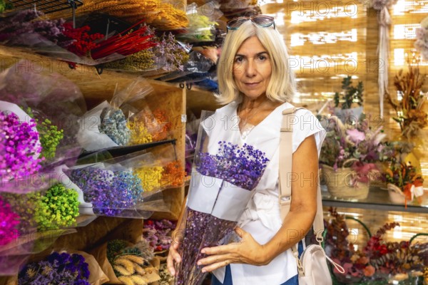 Mature woman with blond hair holds a purple dried flower bouquet inside a colorful market stall filled with handcrafted floral arrangements and natural home decor items for sale