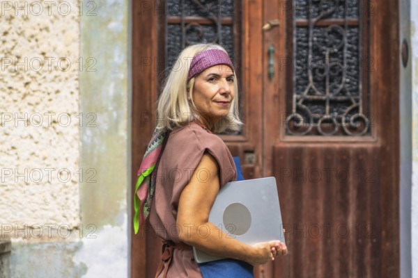Senior woman with gray hair wearing a headband and casual outfit, carrying a laptop and looking back, representing digital nomad lifestyle and modern aging