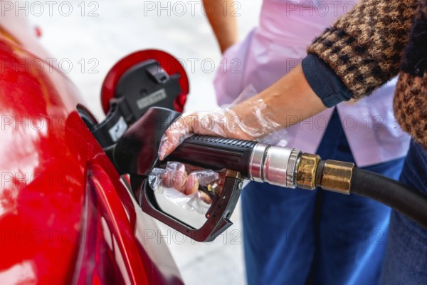 Person is refueling a red car at a gas station, holding a fuel pump nozzle with a protective plastic glove, highlighting the rising costs of gasoline and energy consumption